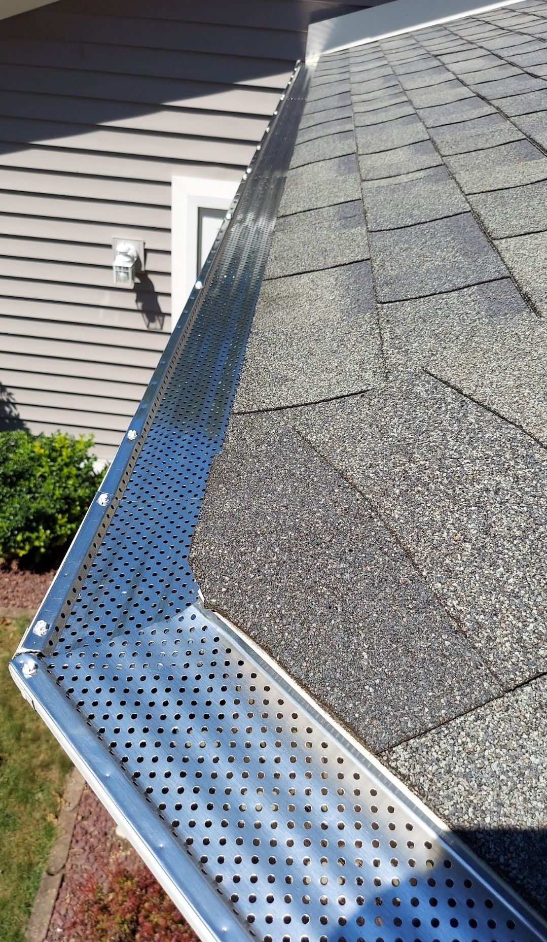 Gutter guard on a gray shingled roof, alongside a light-colored house with some greenery.
