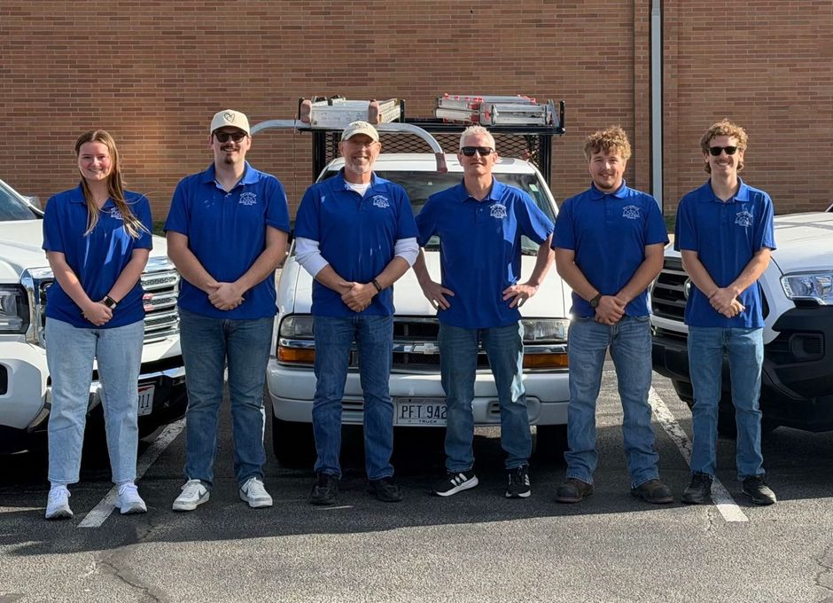 Six people wearing blue polo shirts stand in a row in front of a white service truck parked against a brick wall.
