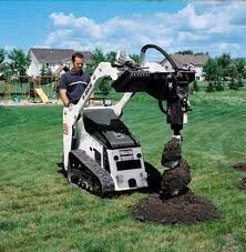 Worker operating a skid-steer excavator digging a hole in a grassy field