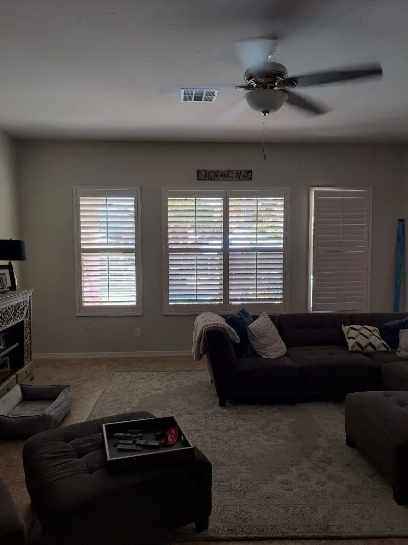 Living room with shutters, sofa, ottoman, and ceiling fan. Soft lighting.