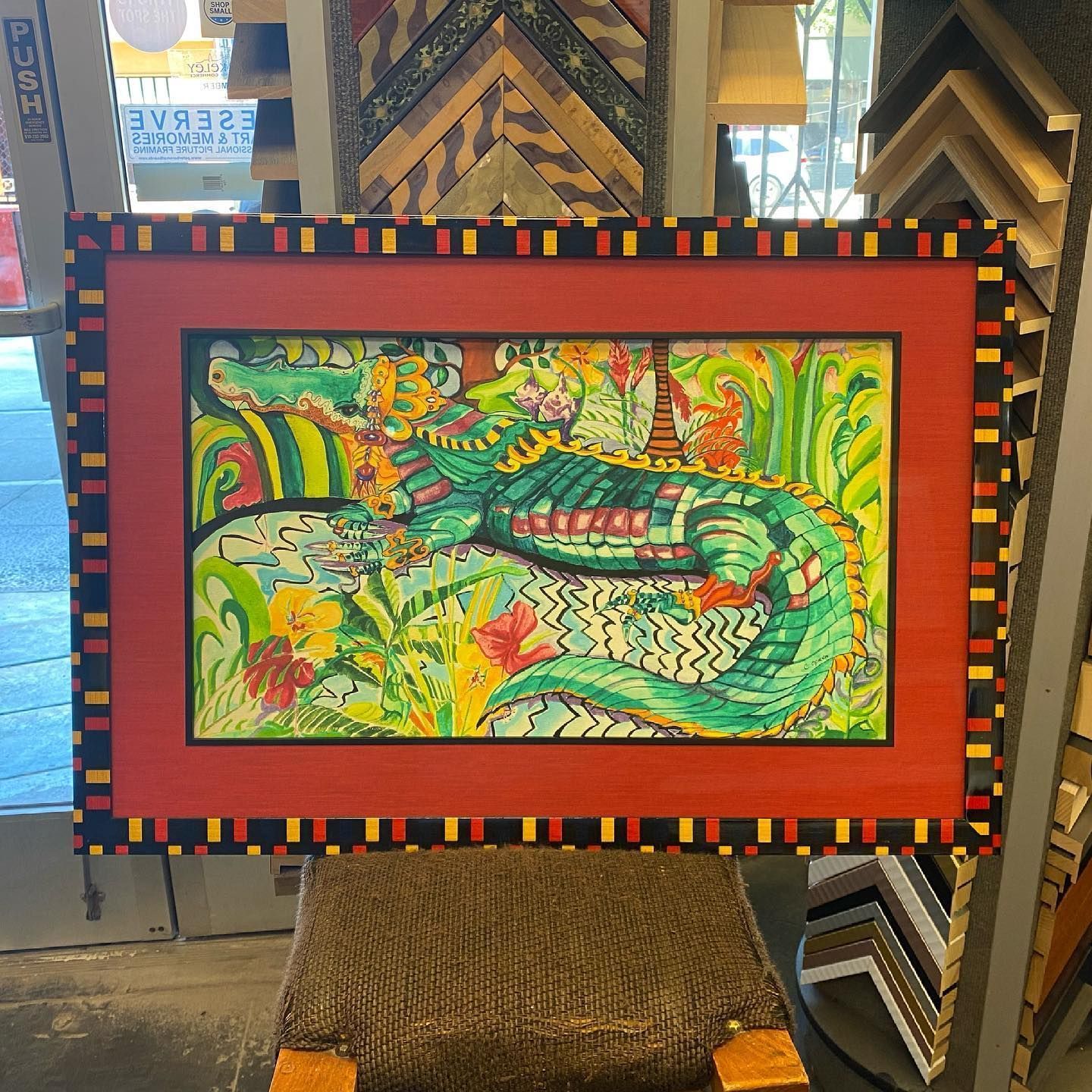 A colorful drawing of an alligator resting in foliage, framed in red with a black and yellow border.