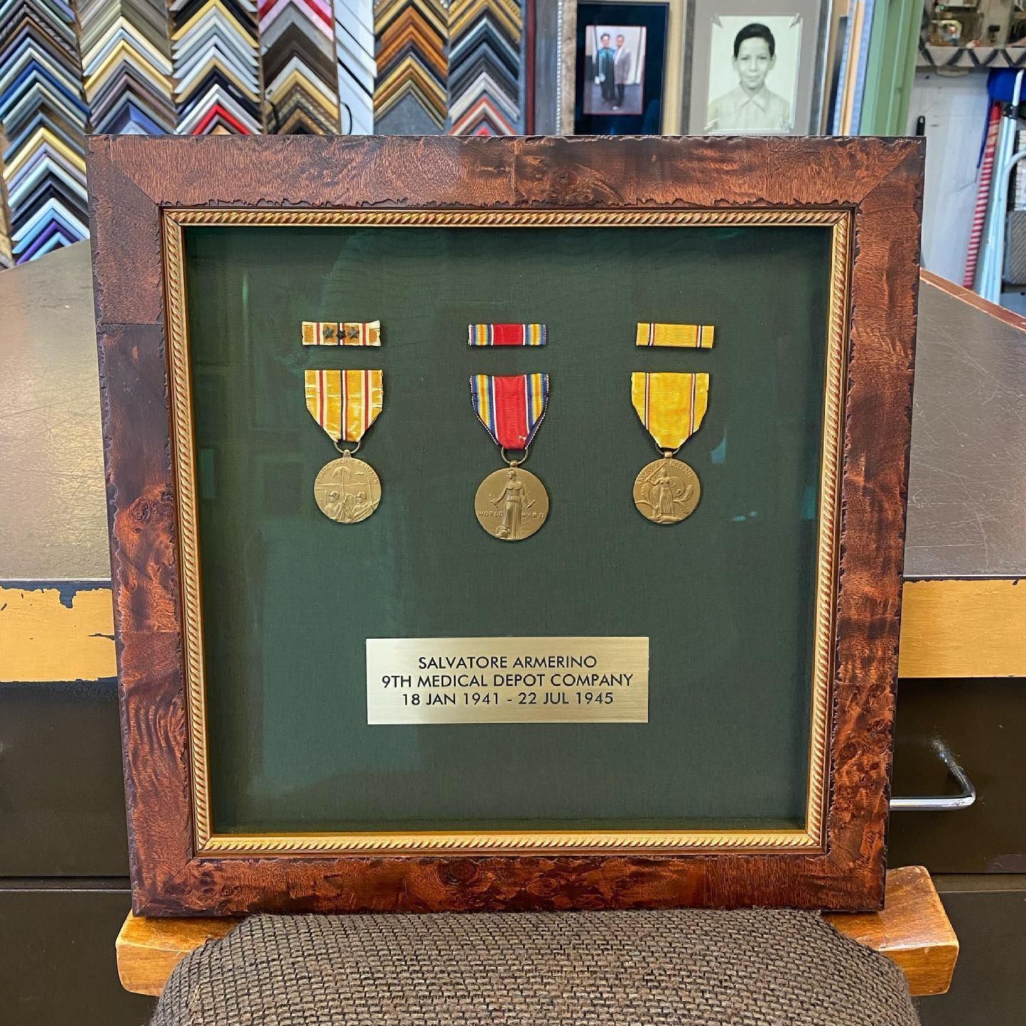 Framed military medals with a nameplate on green background. The frame is brown with a gold inner border.