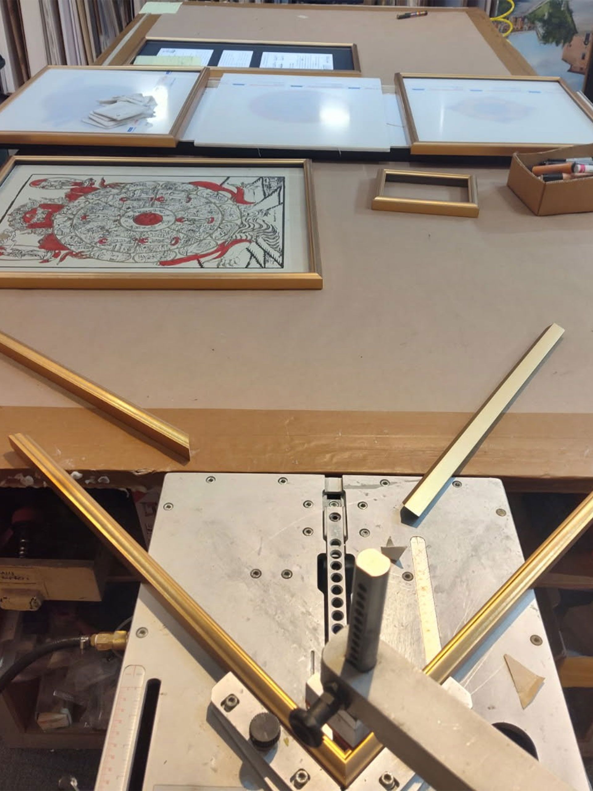 Framing shop with various framed art pieces and a corner joining machine, gold frames and tools.