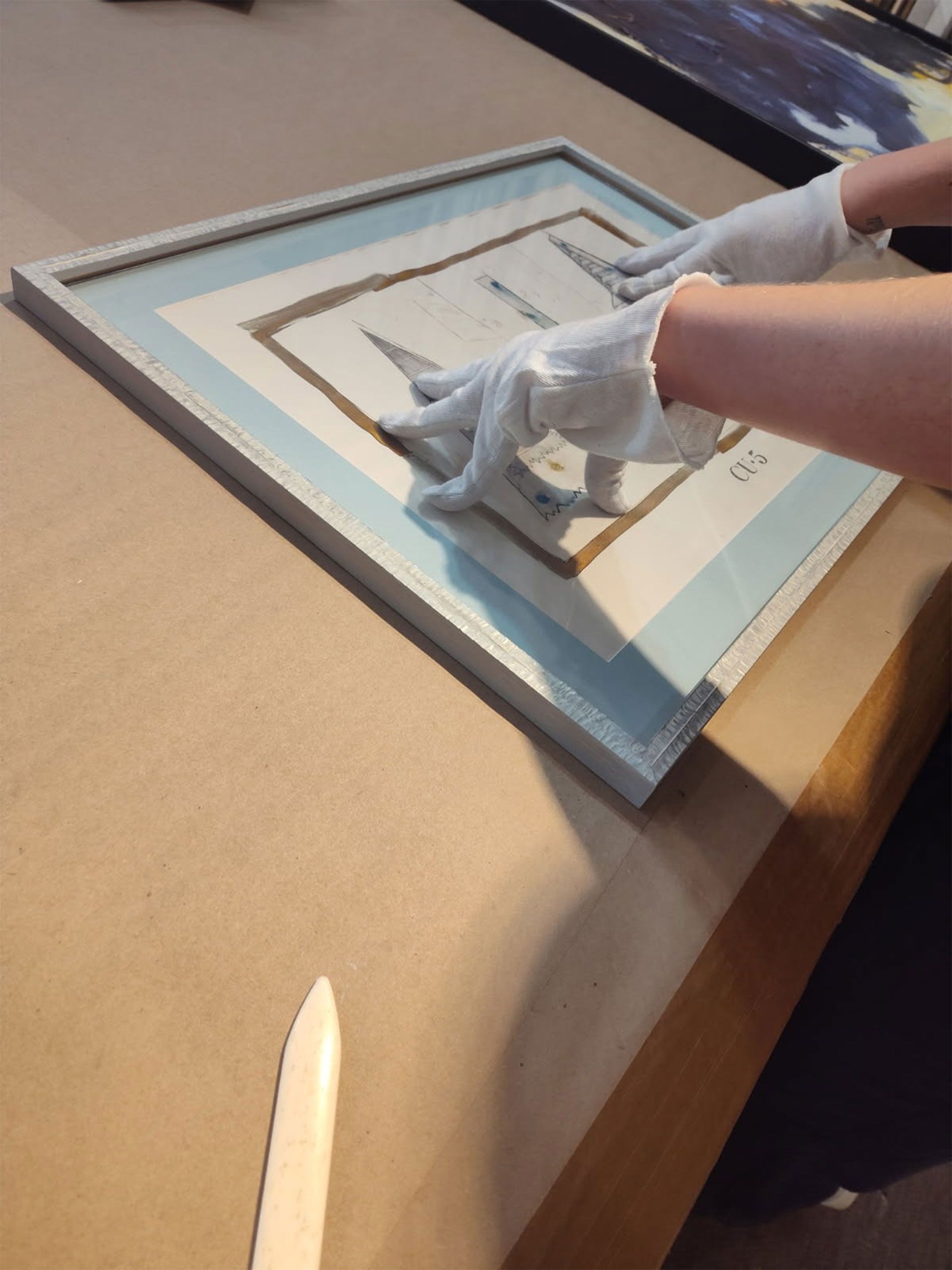 Person wearing gloves handling framed artwork on a table, using a bone folder.