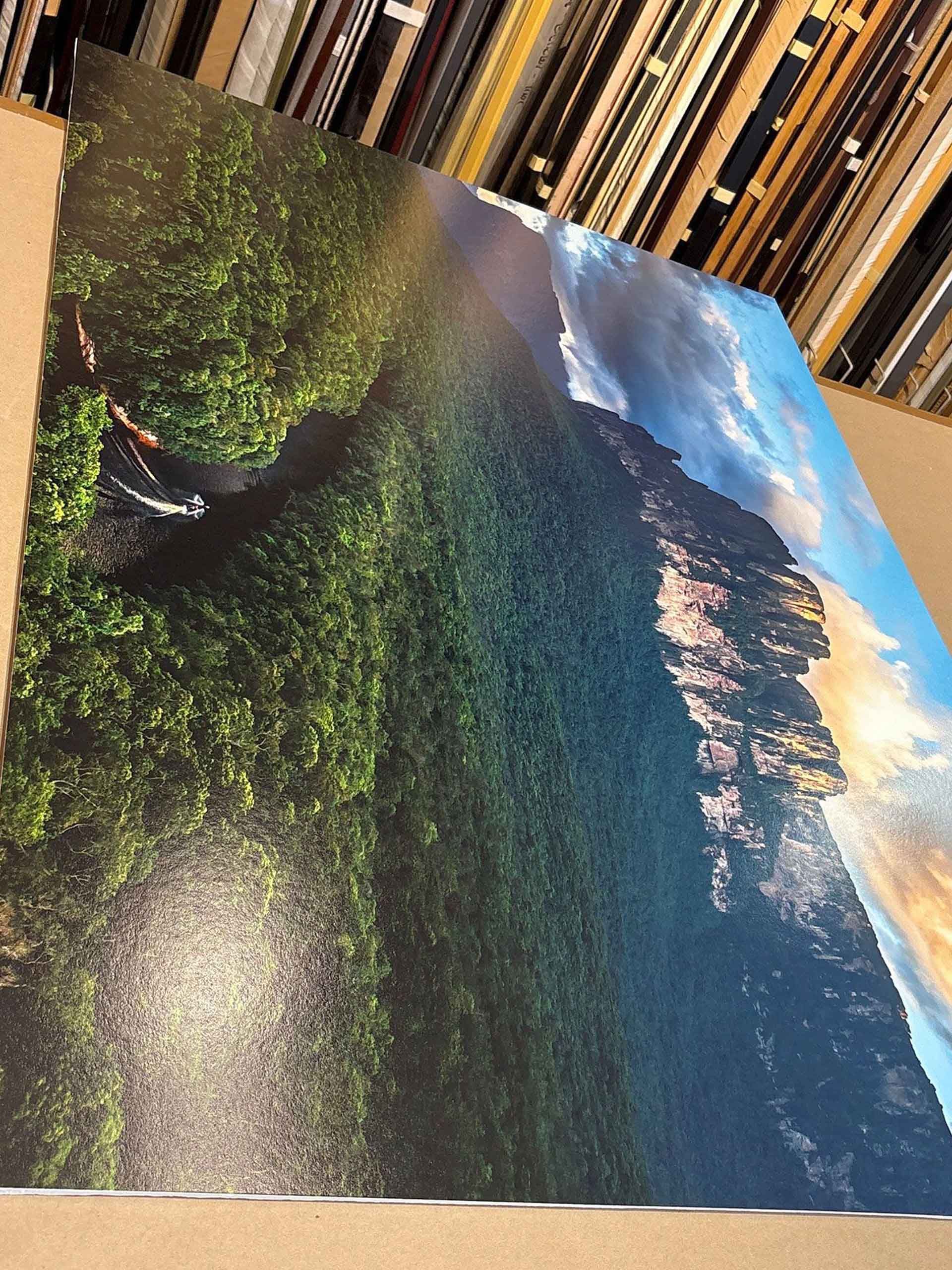 A painting of a lush green forest, waterfall, and rocky cliff, with a blue sky