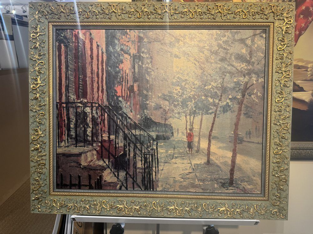 Painting of a snow-covered street scene with a red building and a person walking down a path. The painting is in a gold ornate frame.