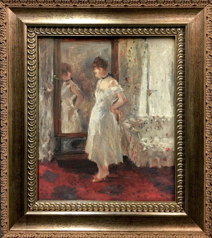 Woman in white dress looking in a mirror; standing on a red rug in front of a window. Oil painting in a gold frame.