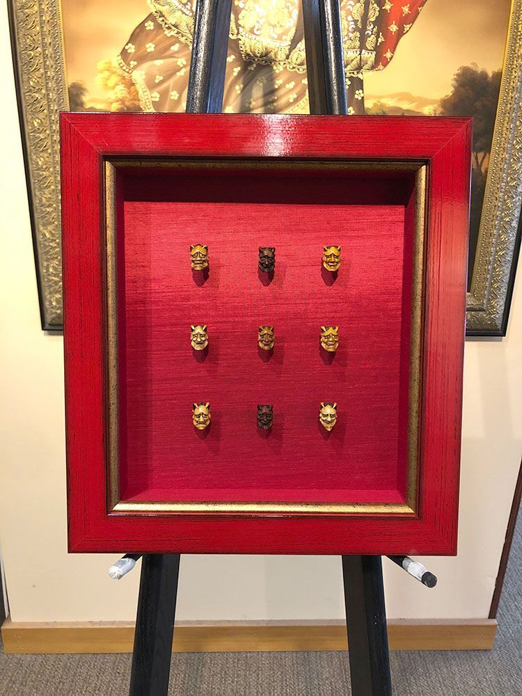 Nine small golden masks displayed in a red framed shadow box on an easel