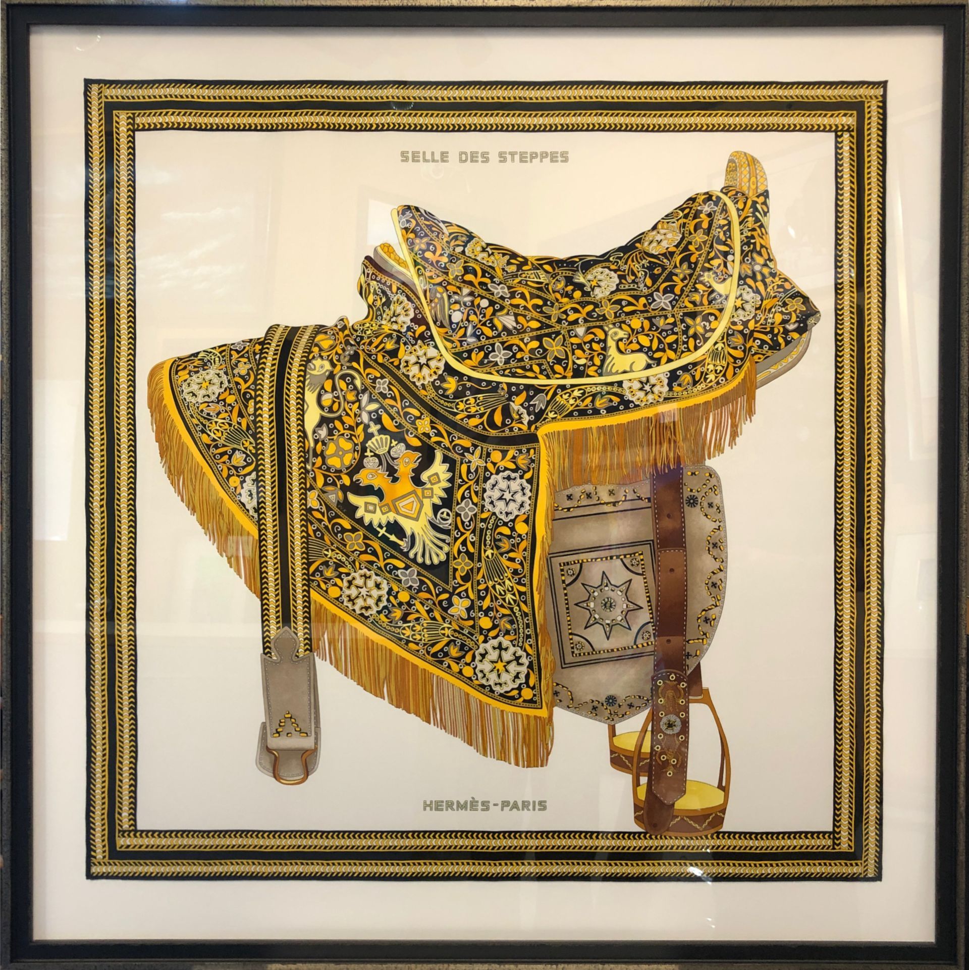 Framed ornate black-and-gold embroidered saddle with tassels, displayed against a light background