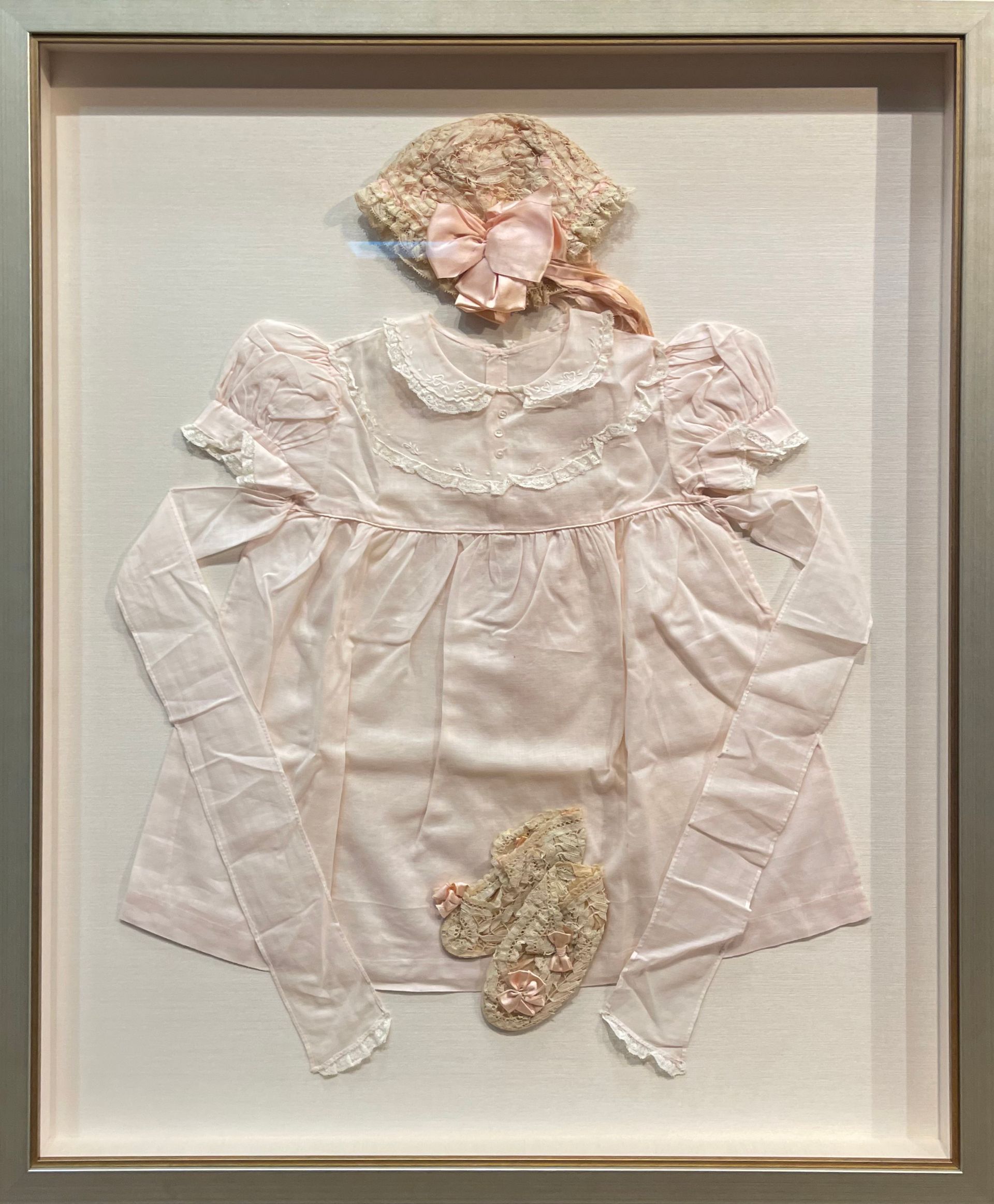 Framed white ruffled dress with floral bonnet and matching shoes displayed on a cream background