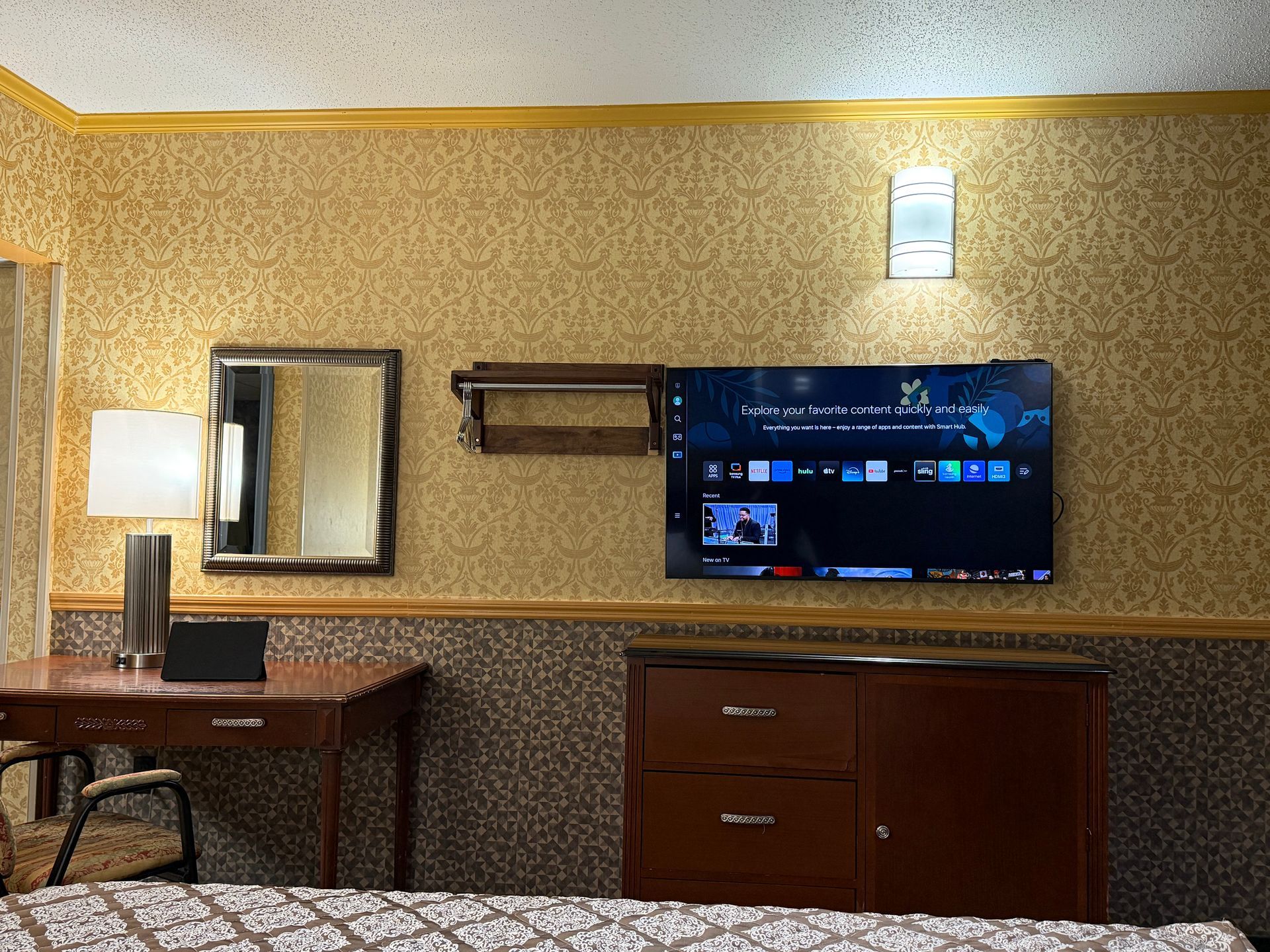 Hotel room with desk, mirror, TV, and dresser. Gold patterned wallpaper.