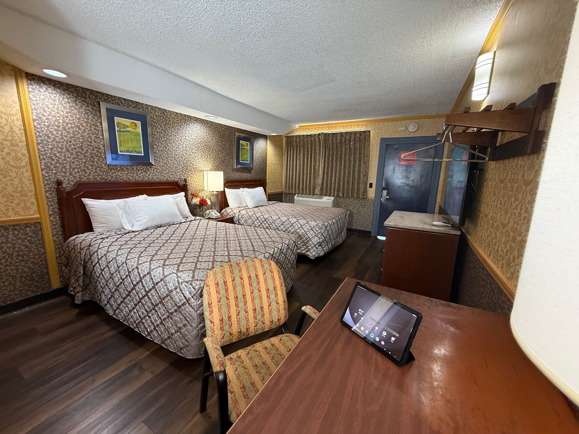 Hotel room with two beds, a desk, chair, and a wardrobe. Brown and gold color scheme.