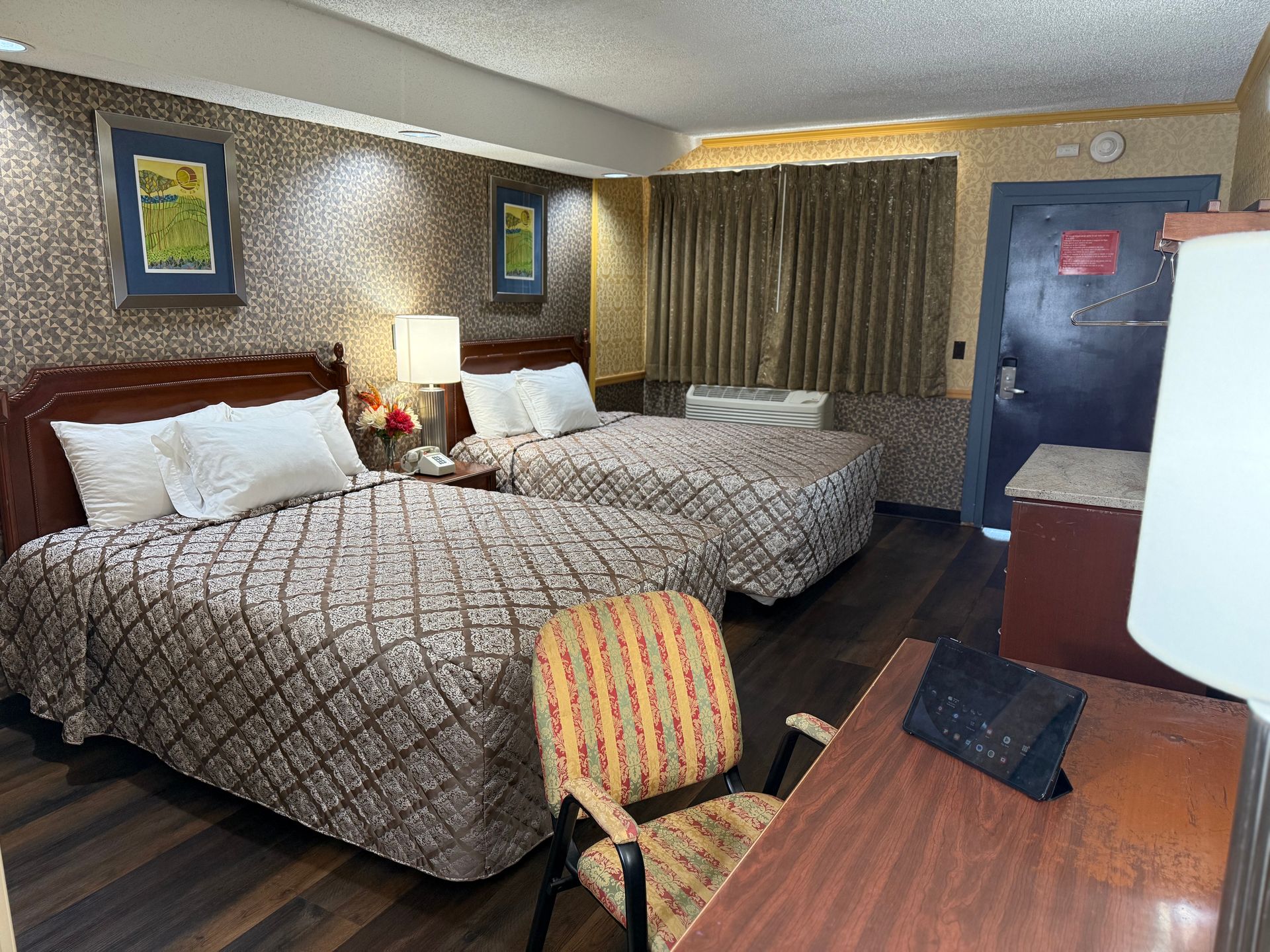 Hotel room with two beds, desk, and chair. Brown patterned bedspreads, patterned wallpaper, and closed curtains.