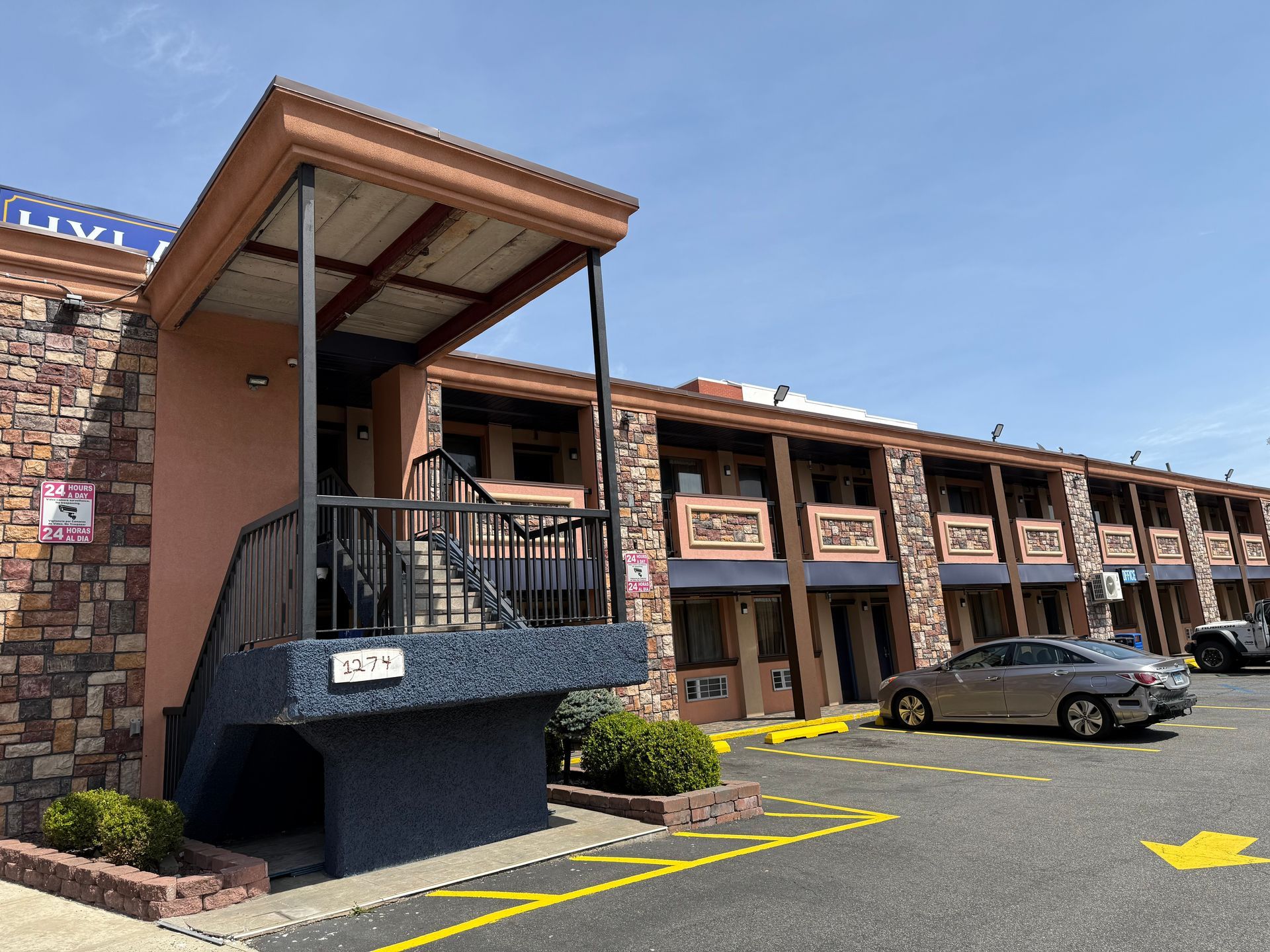 Motel exterior with stairwell, brick facade, parked cars, and blue sky.