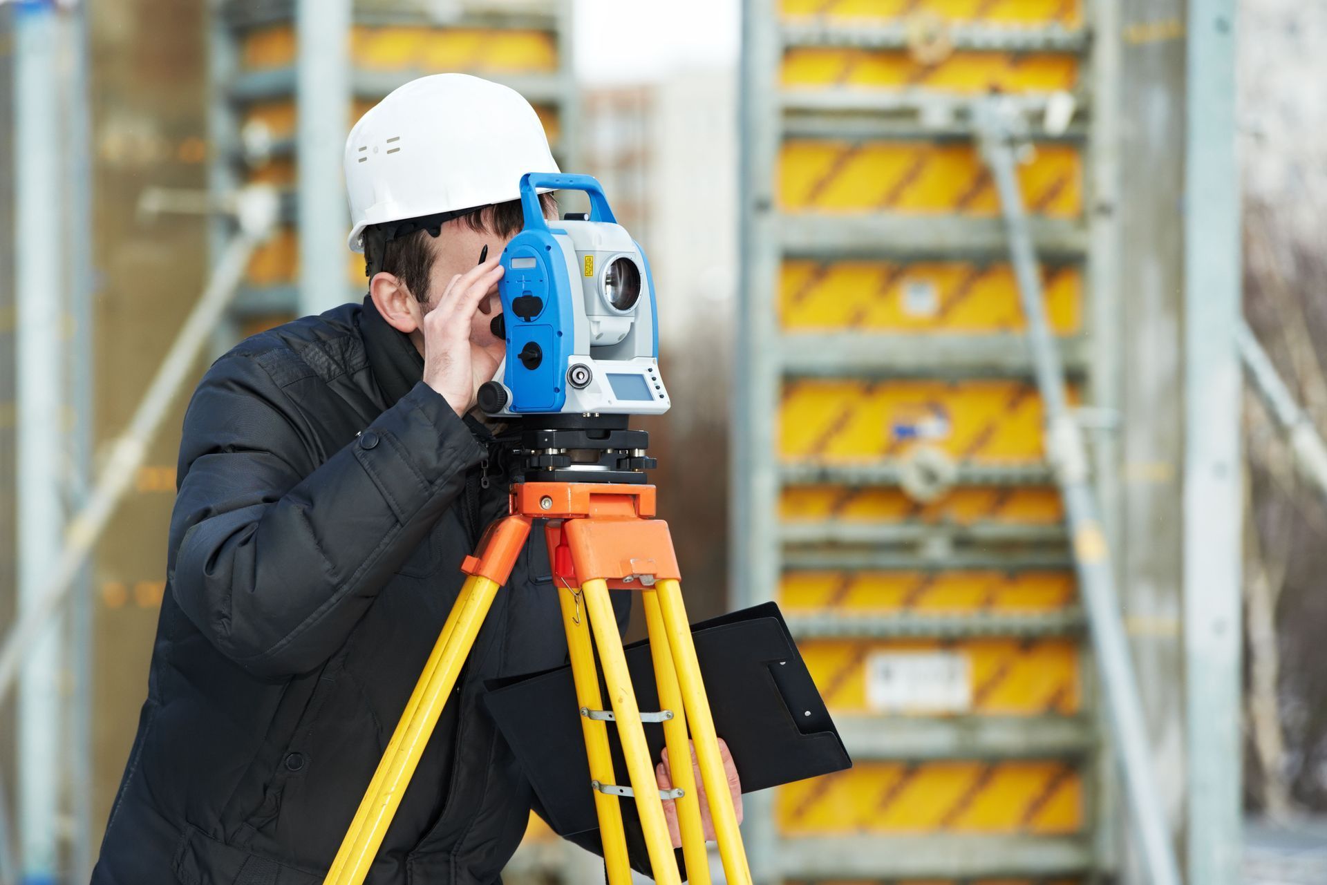 Surveyor using a total station on a tripod at a construction site, wearing a white hard hat.