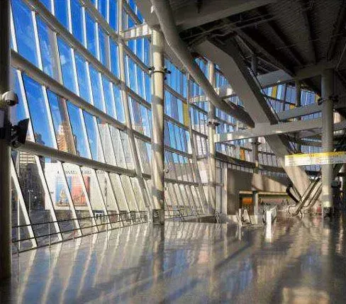 Modern glass-and-steel atrium with sunlight, reflective floor, and diagonal support beams