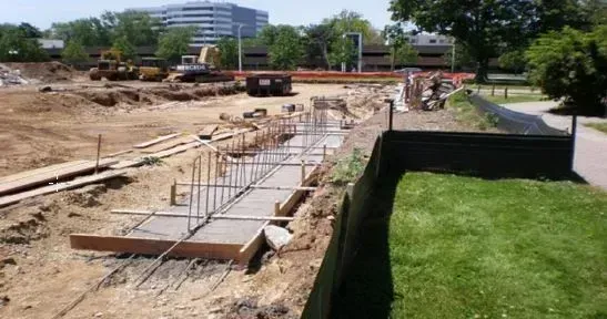 Construction site with concrete foundation forms and rebar beside a grassy lawn under a sunny sky