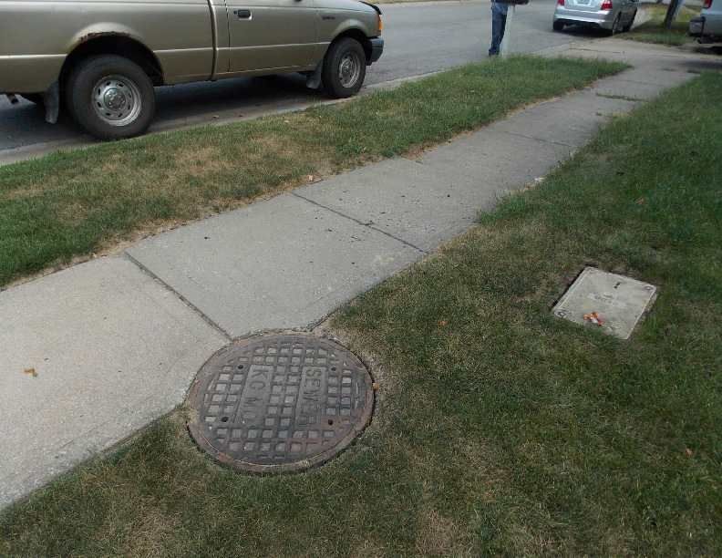 Sidewalk with a manhole cover beside parked cars on a grassy residential street.