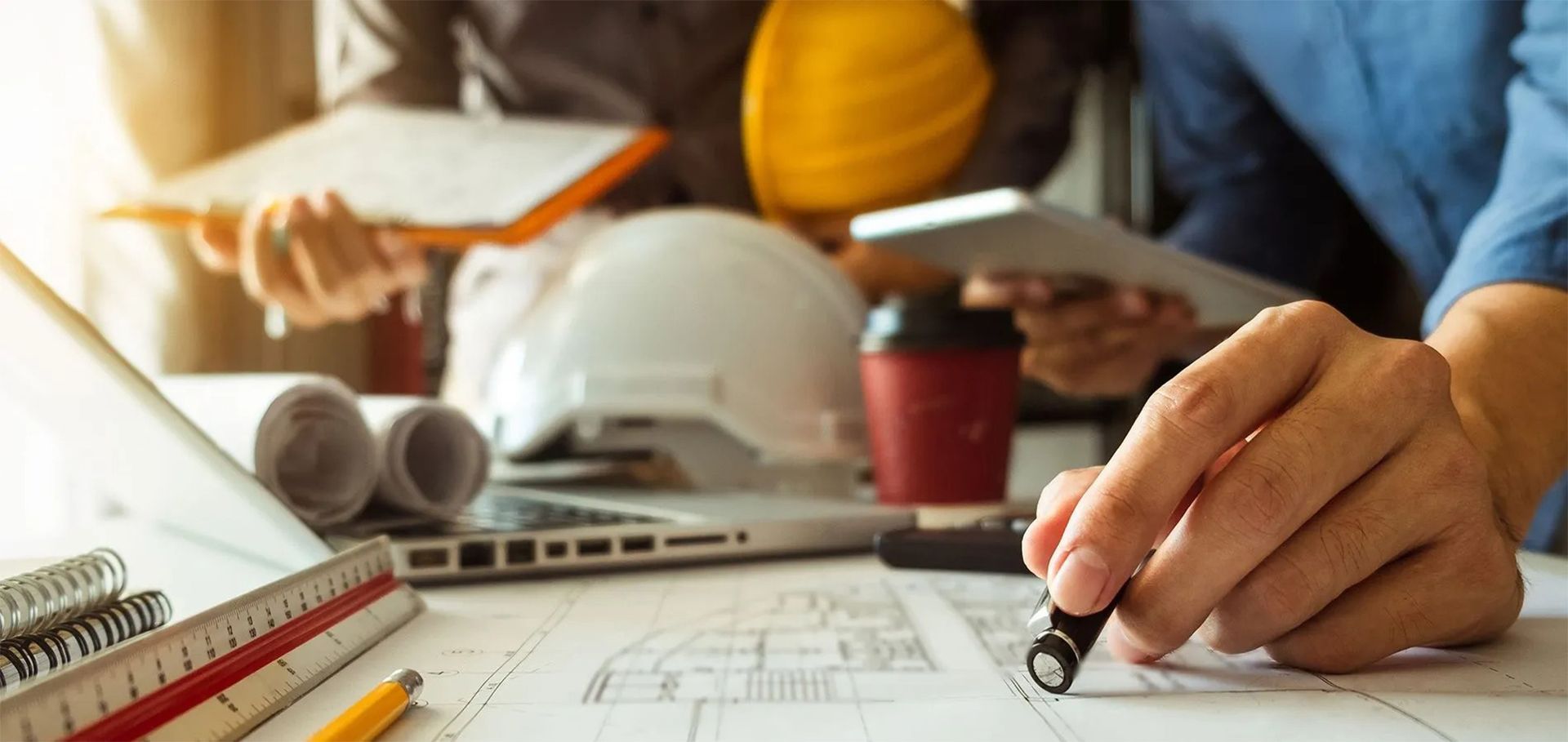 Architects reviewing blueprints at a desk with a hard hat, pencil, and coffee cup