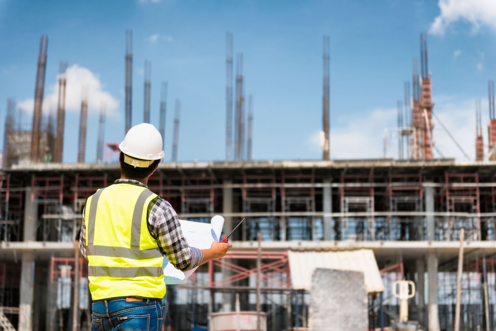 Construction worker in white hard hat and yellow vest surveying a building site under construction
