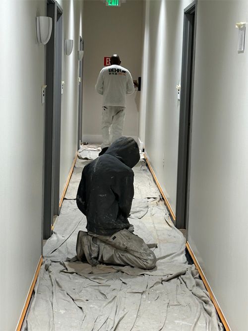 A man is kneeling down in a hallway while another man walks behind him.