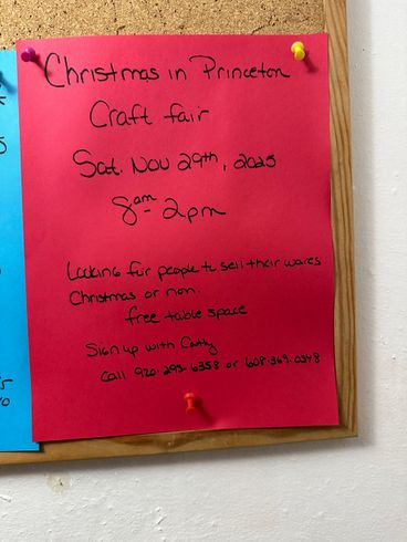 Red paper on a bulletin board advertises a Christmas craft fair in Princeton, Saturday, November 29th, 8 am-2 pm. Free vendor space available.