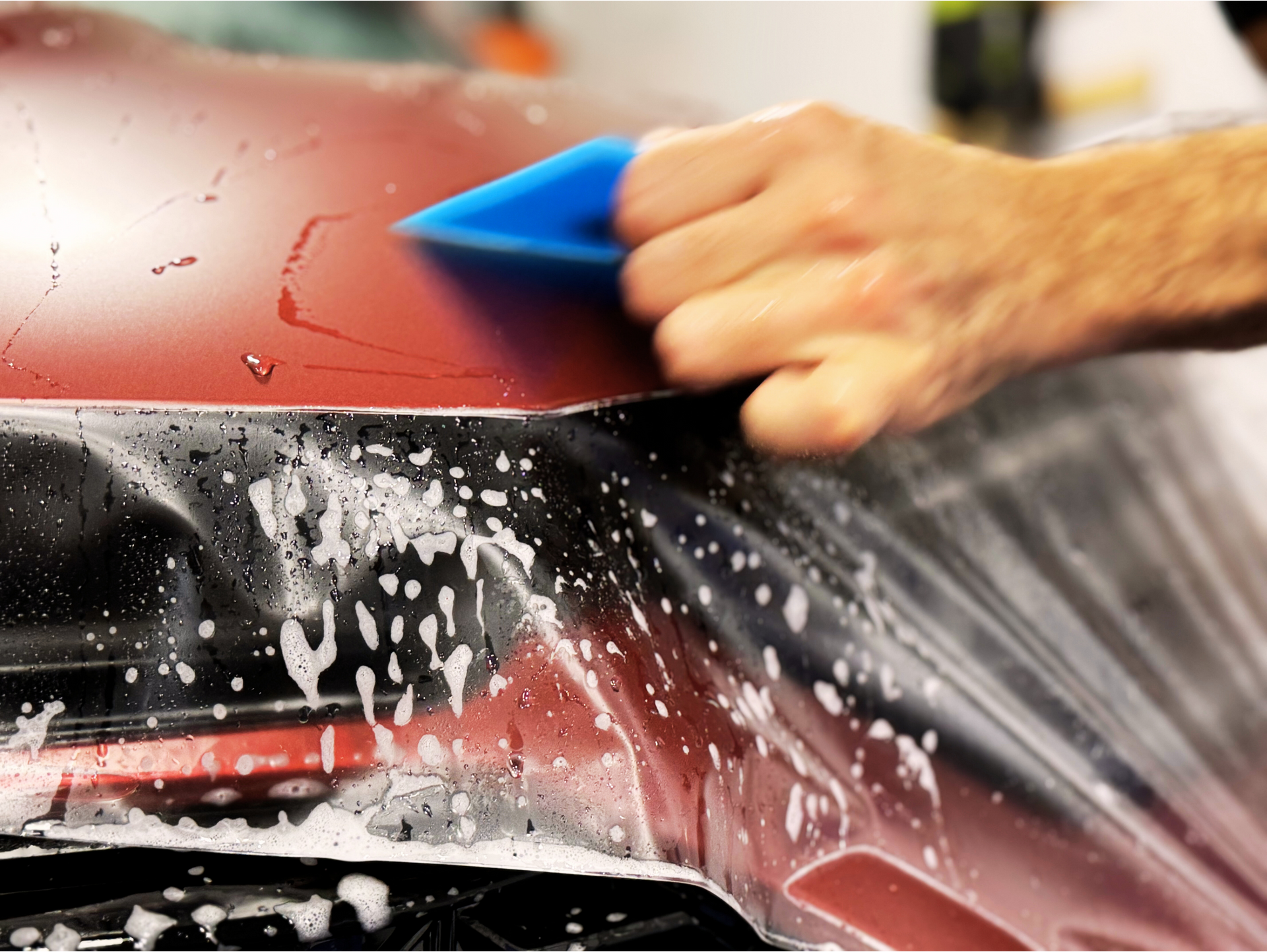 Person applying protective film to a red car hood with a blue squeegee.