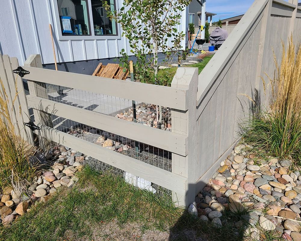 A wooden fence is surrounded by rocks and grass in front of a house.