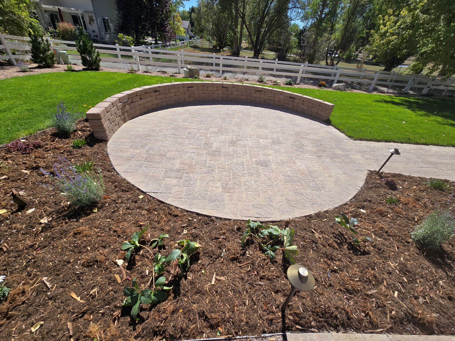A circular patio in the middle of a garden with a white fence in the background
