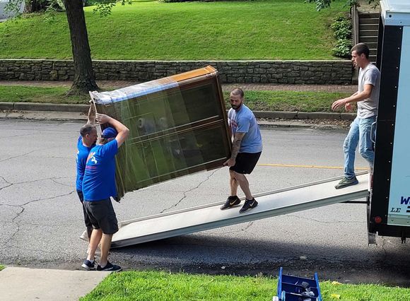 Four people moving a large, brown cabinet onto a truck via a ramp on a sunny day.