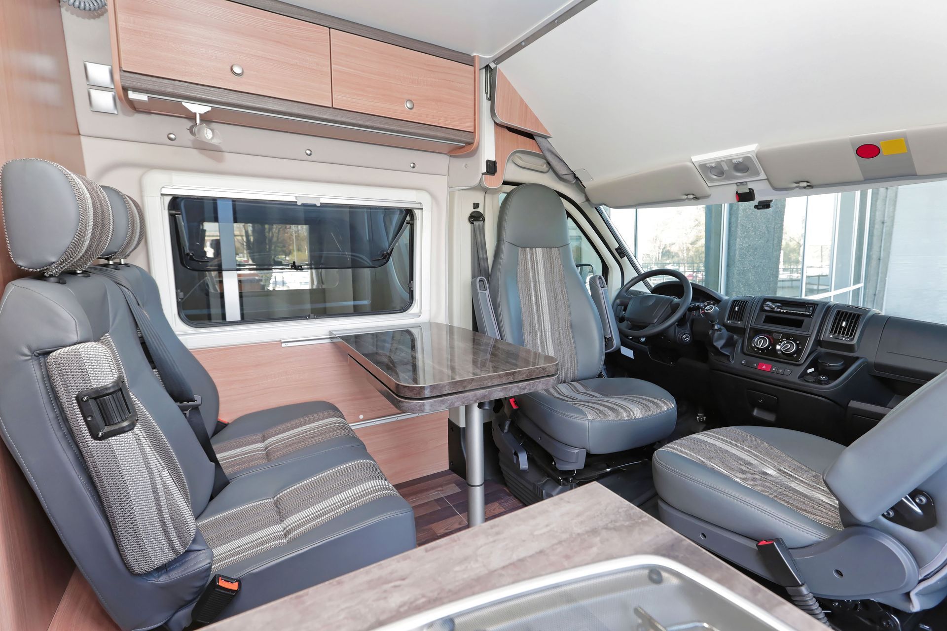Compact RV interior with dinette, front cab seats, and overhead storage in gray and wood tones
