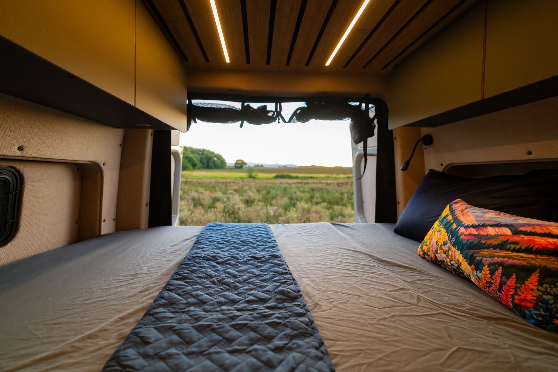 Interior of a camper van with a bed facing an open rear hatch and a grassy field outside