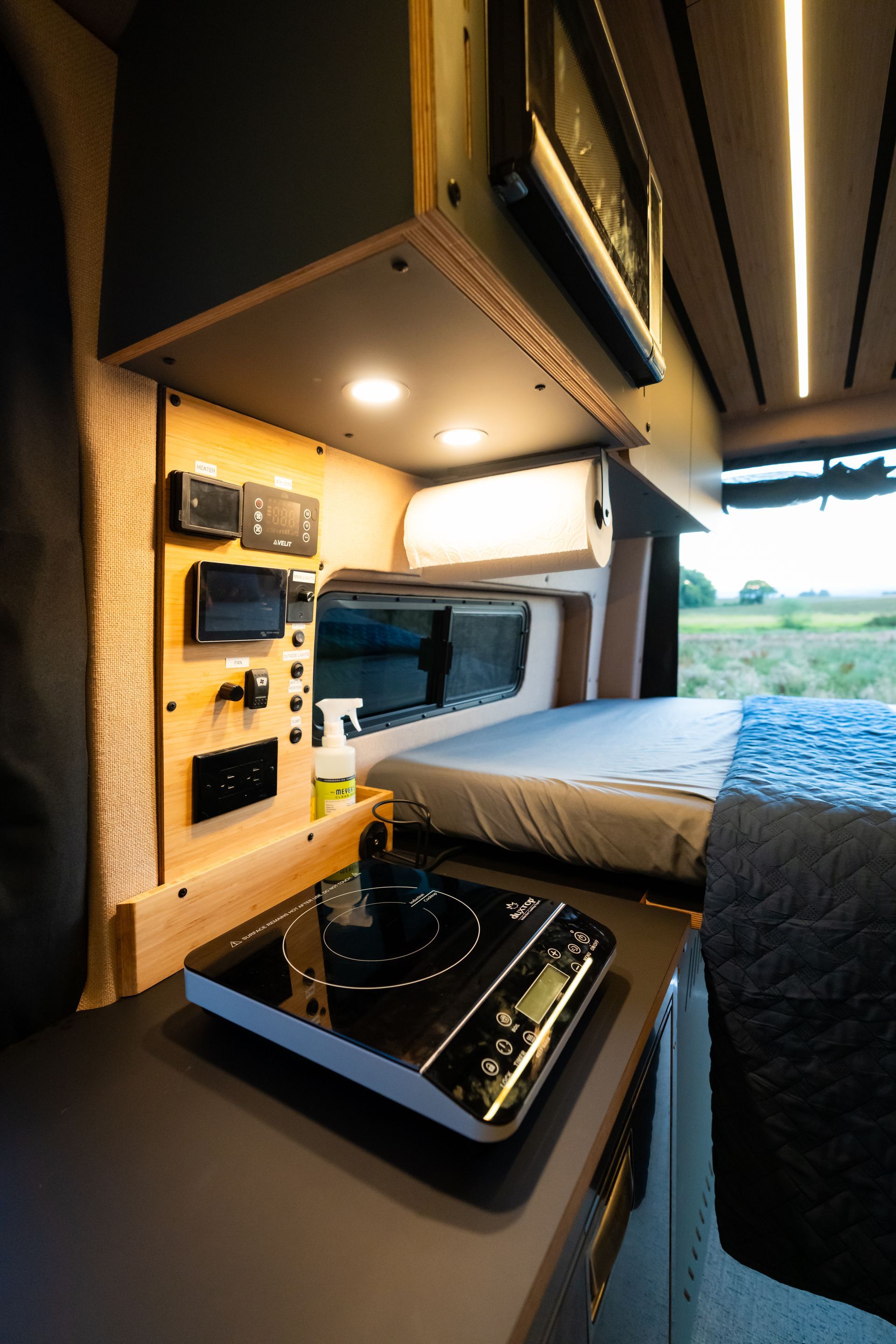 Compact camper van interior with bed, overhead storage, desk, and a small kitchenette by the window