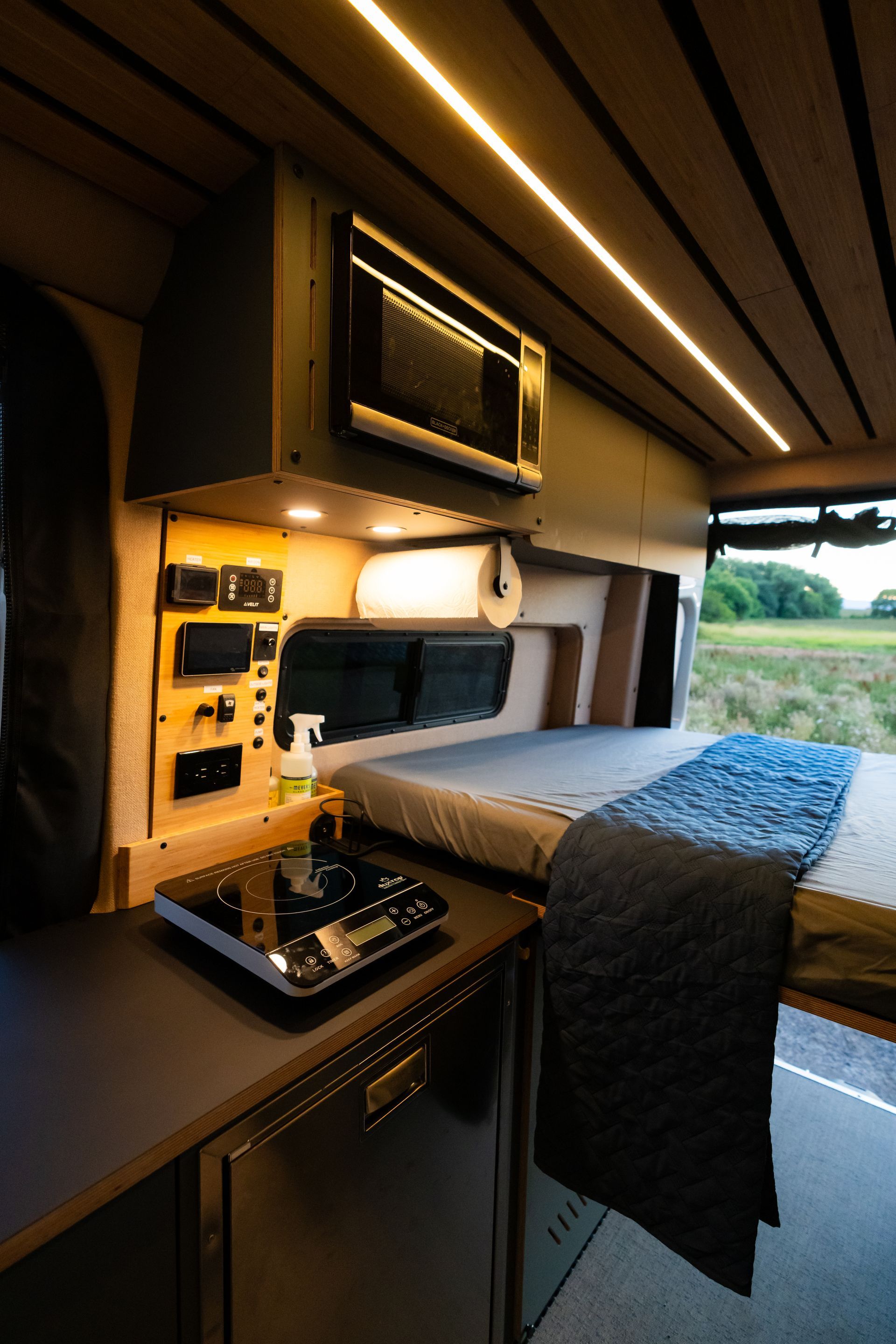 Compact camper van interior with bed, overhead storage, and open side door overlooking a field