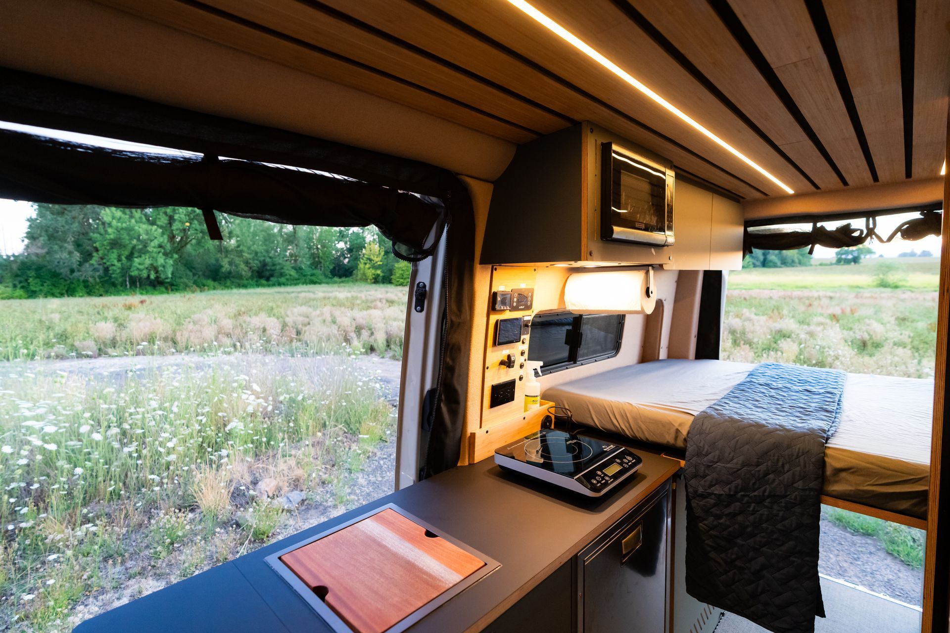 Cozy camper van interior with bed, desk, and open side door overlooking a grassy field