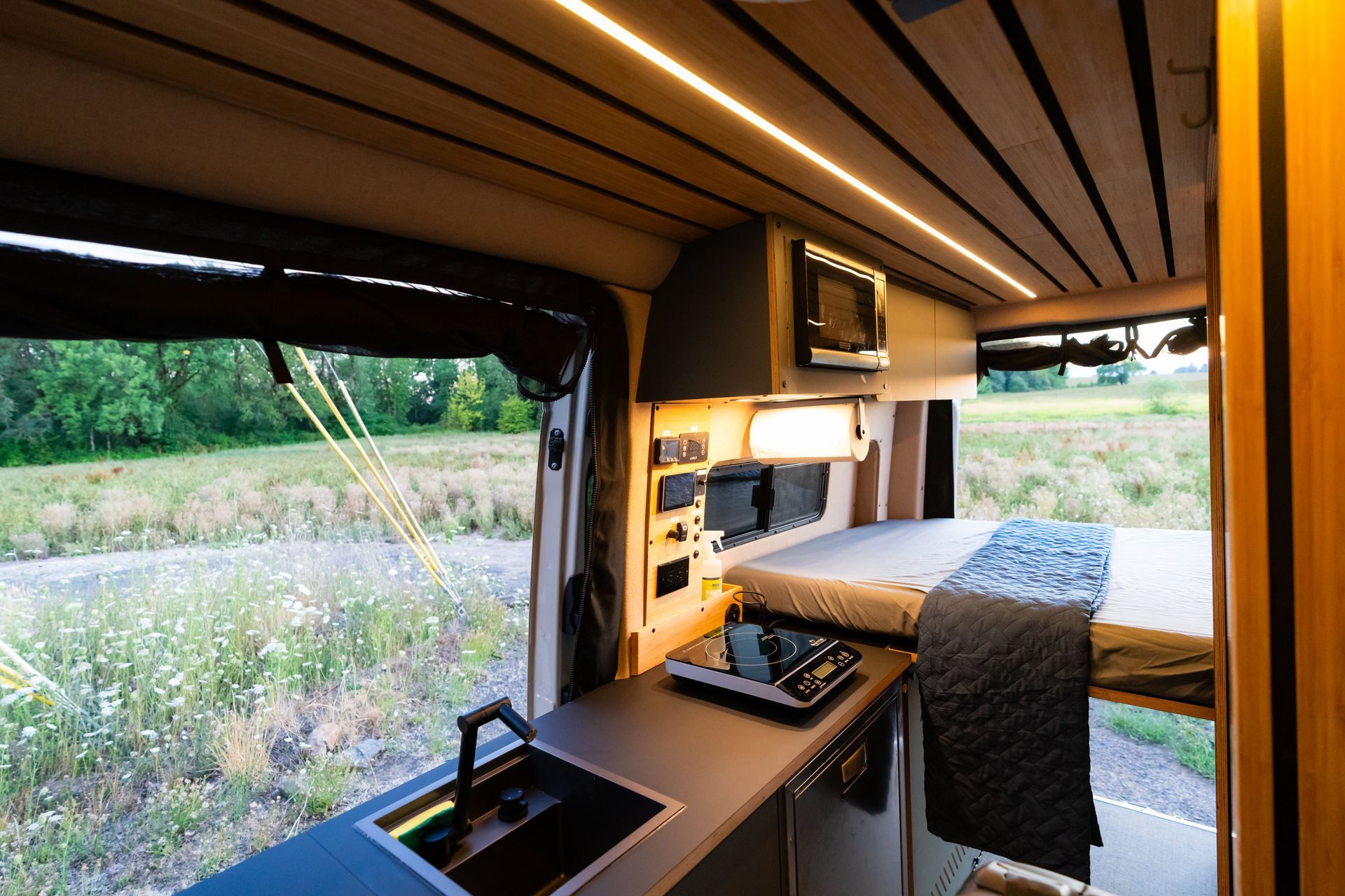 Compact camper van interior with a bed, desk, and large open side window overlooking greenery