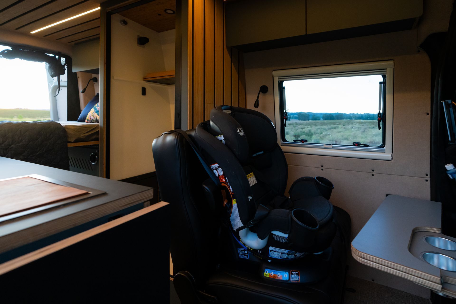 Interior of a camper van with a child car seat, window view of open countryside, and a compact table.