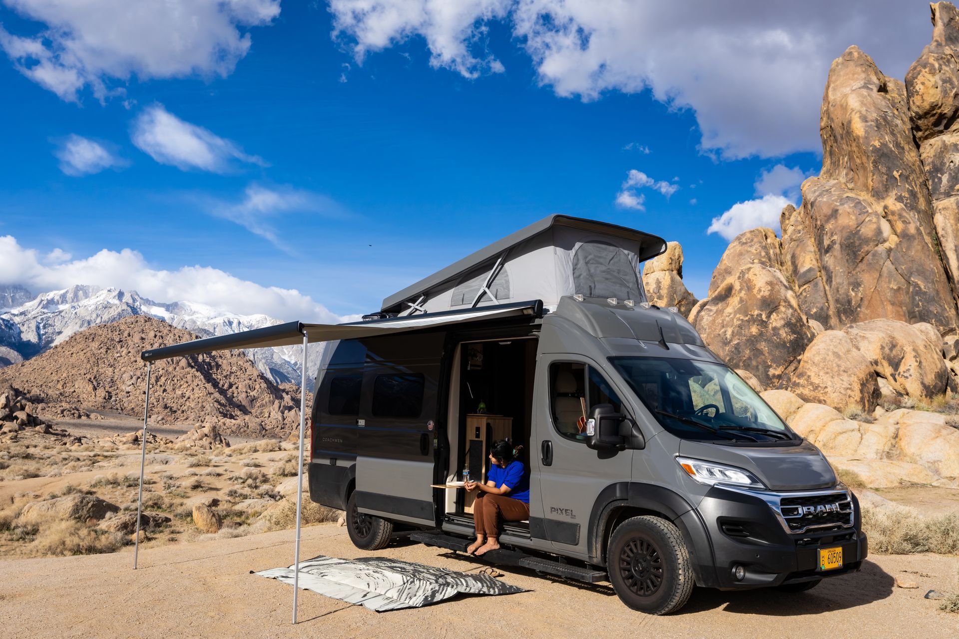 Gray camper van parked in a rocky desert under a blue sky, with awning extended and a chair outside.