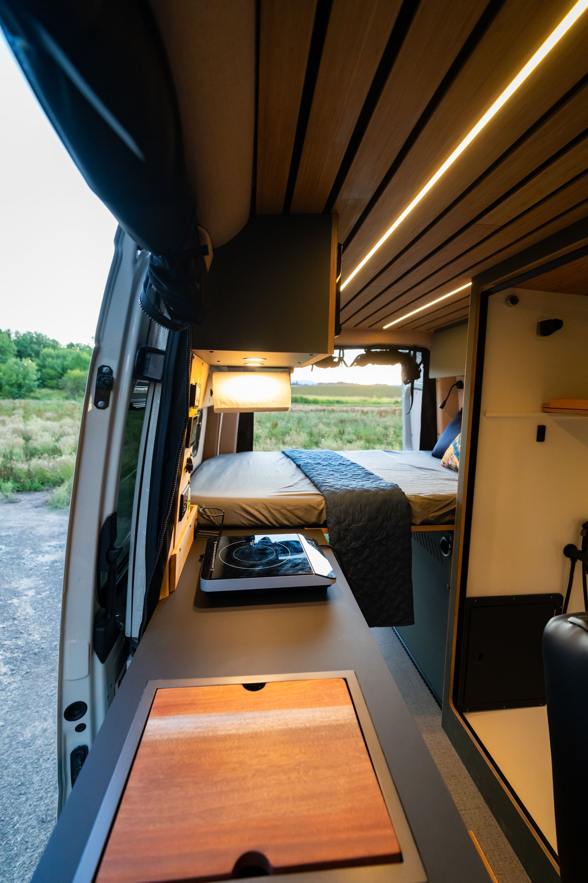 Compact camper van interior with a bed, warm lighting, wood ceiling, and open rear doors looking out to greenery