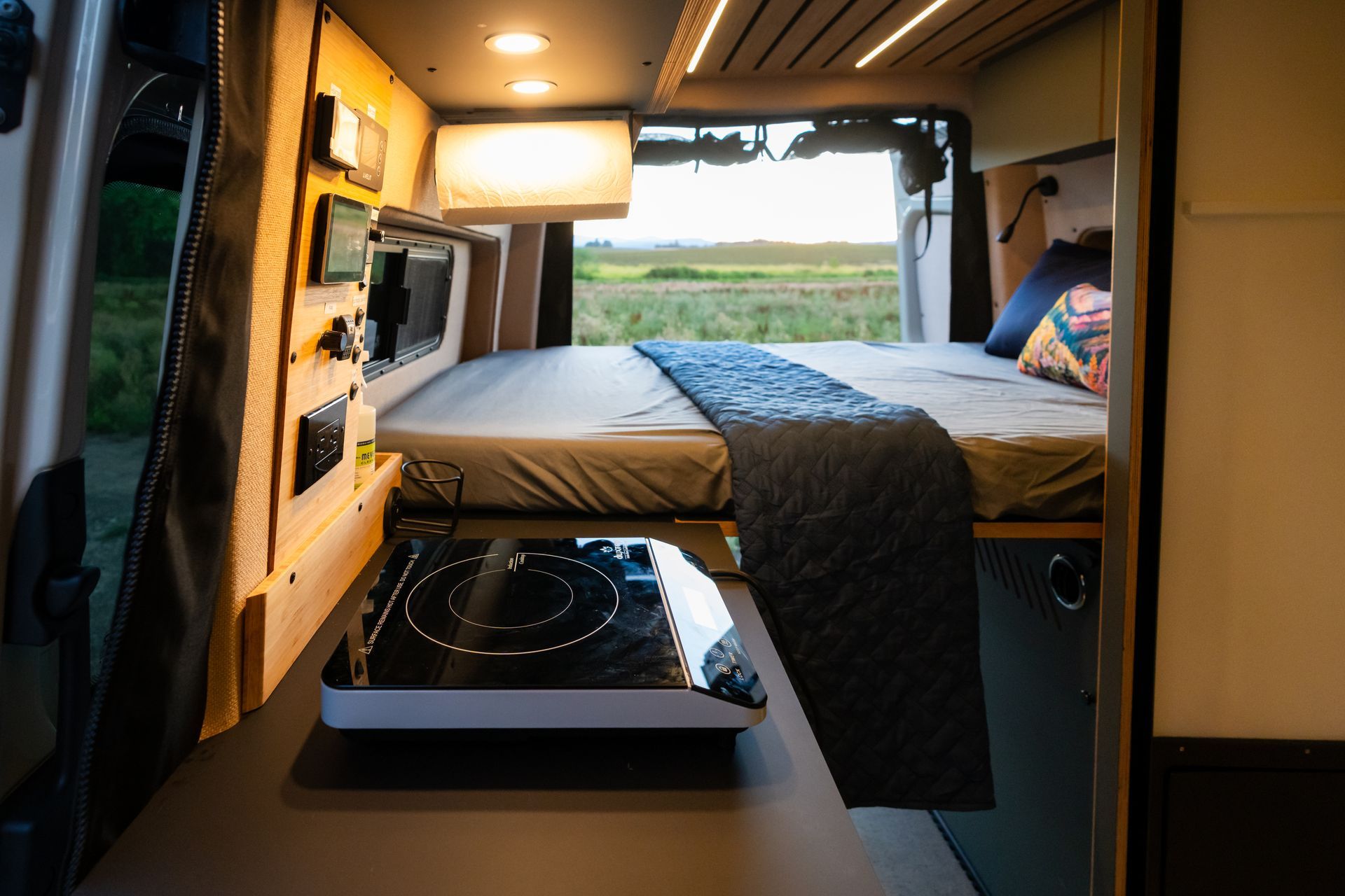 Compact camper van interior with double bed, small kitchen, and open rear doors overlooking a green field.