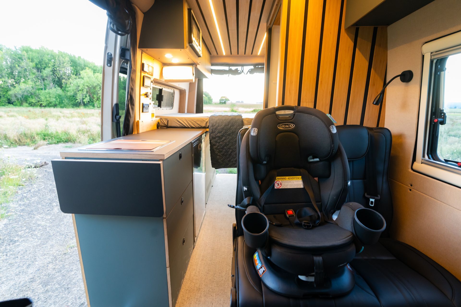 Interior of a small camper van with a black child car seat and wood-paneled walls, parked by greenery