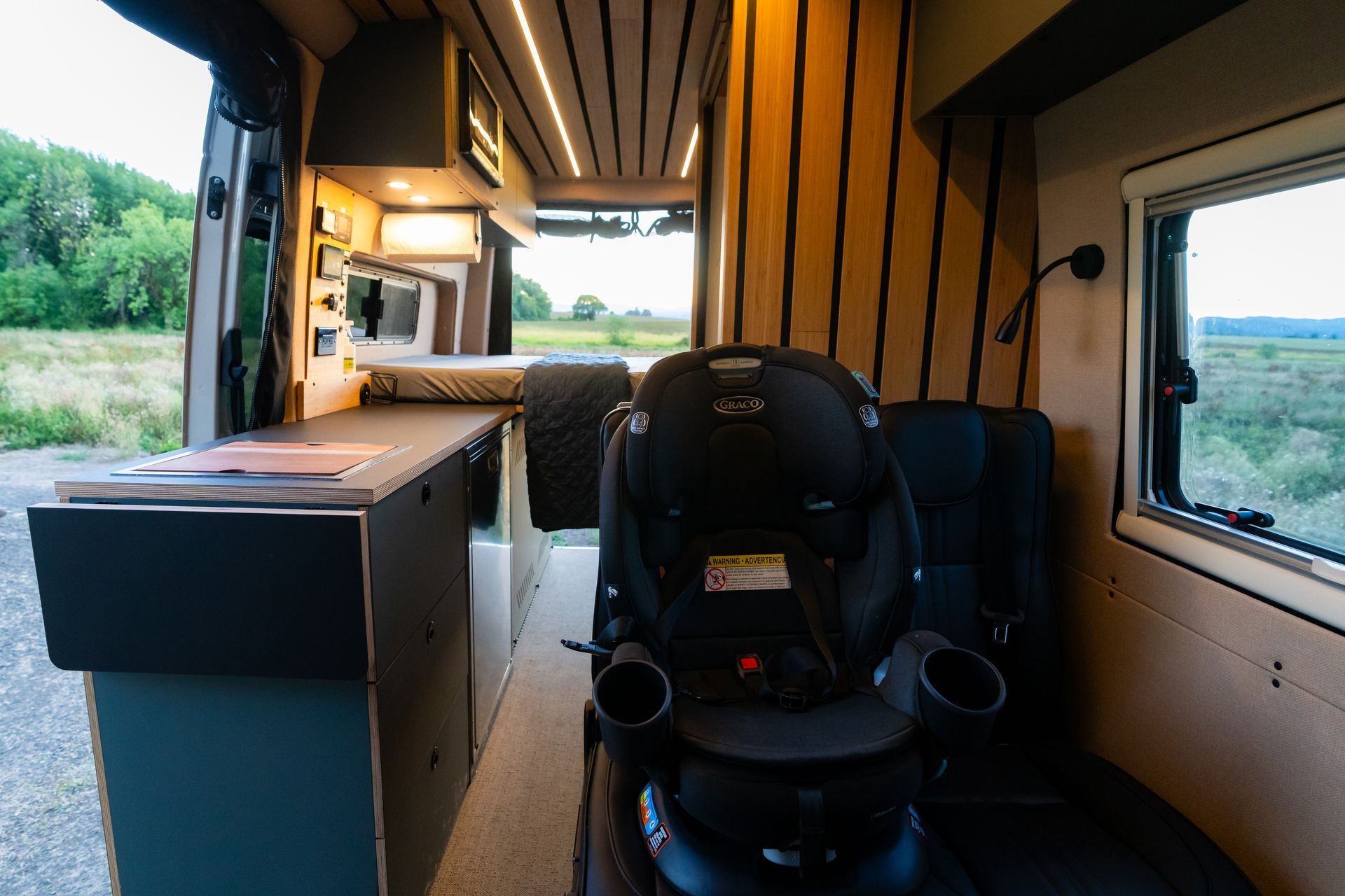 Inside a modern bus, a child car seat faces forward beside large windows and a view of greenery.