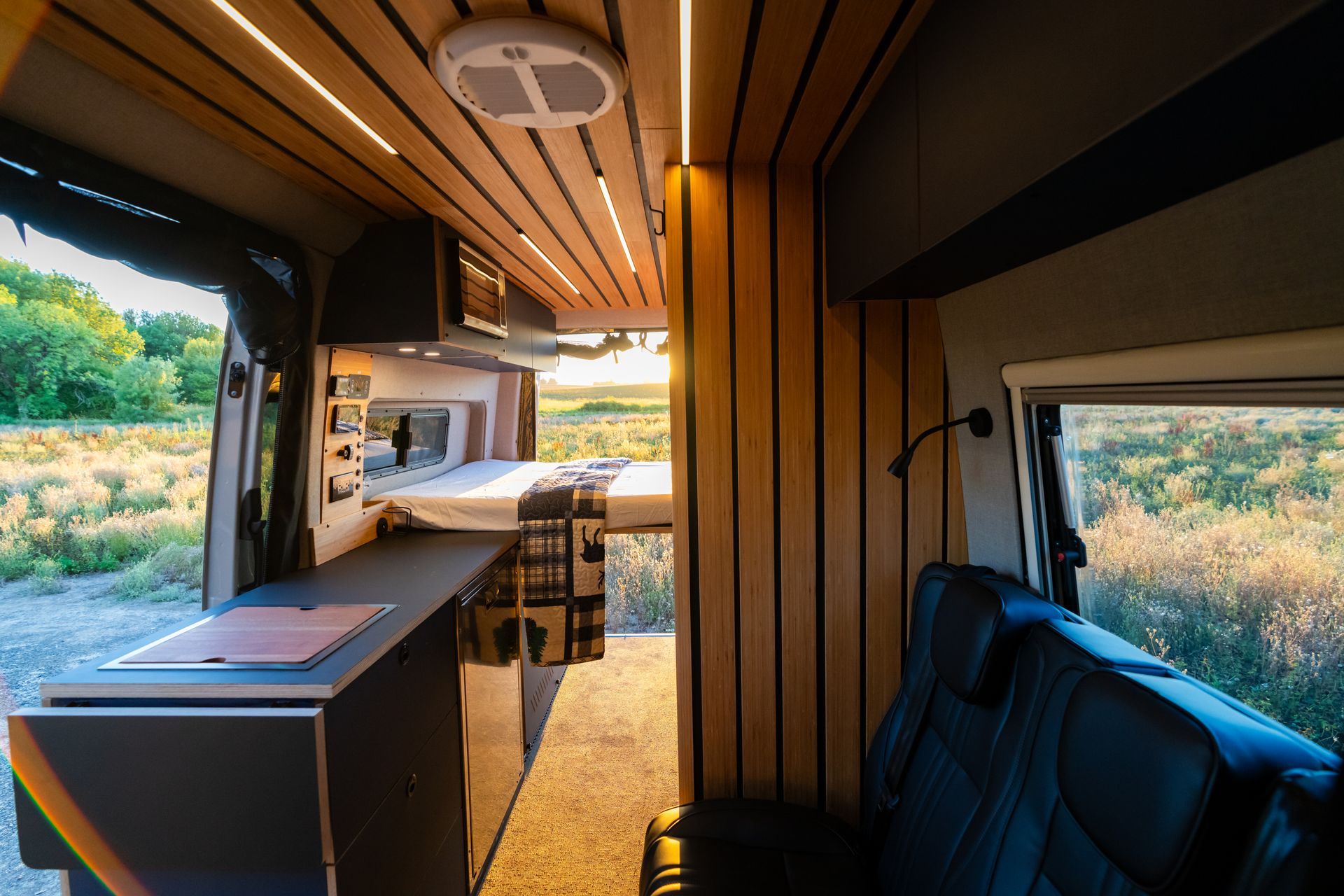 Modern camper van interior with wood paneling, sink, table, and open view to grassy landscape