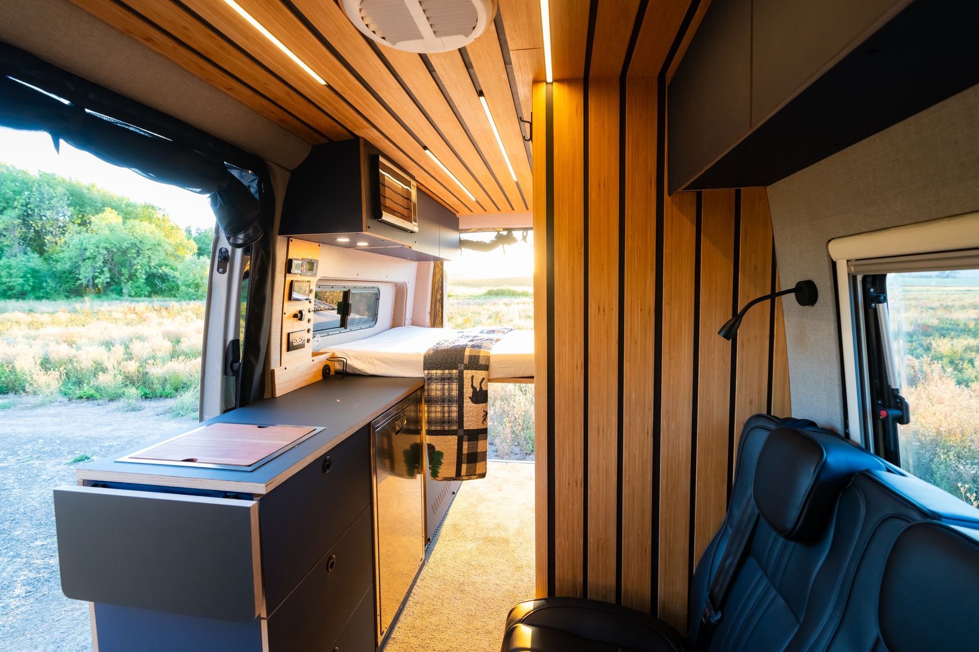 Interior of a camper van with wood paneling, a built-in countertop, and an open side door overlooking outdoors