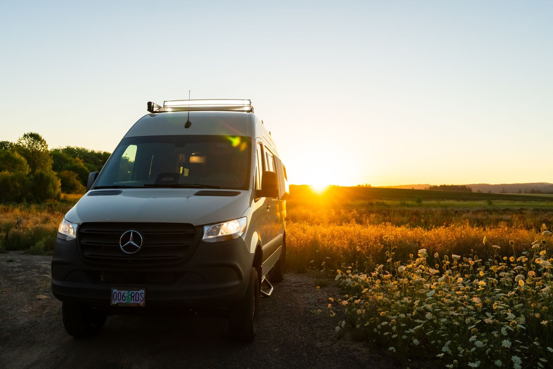 White van on a dirt road at sunset beside a field of wildflowers