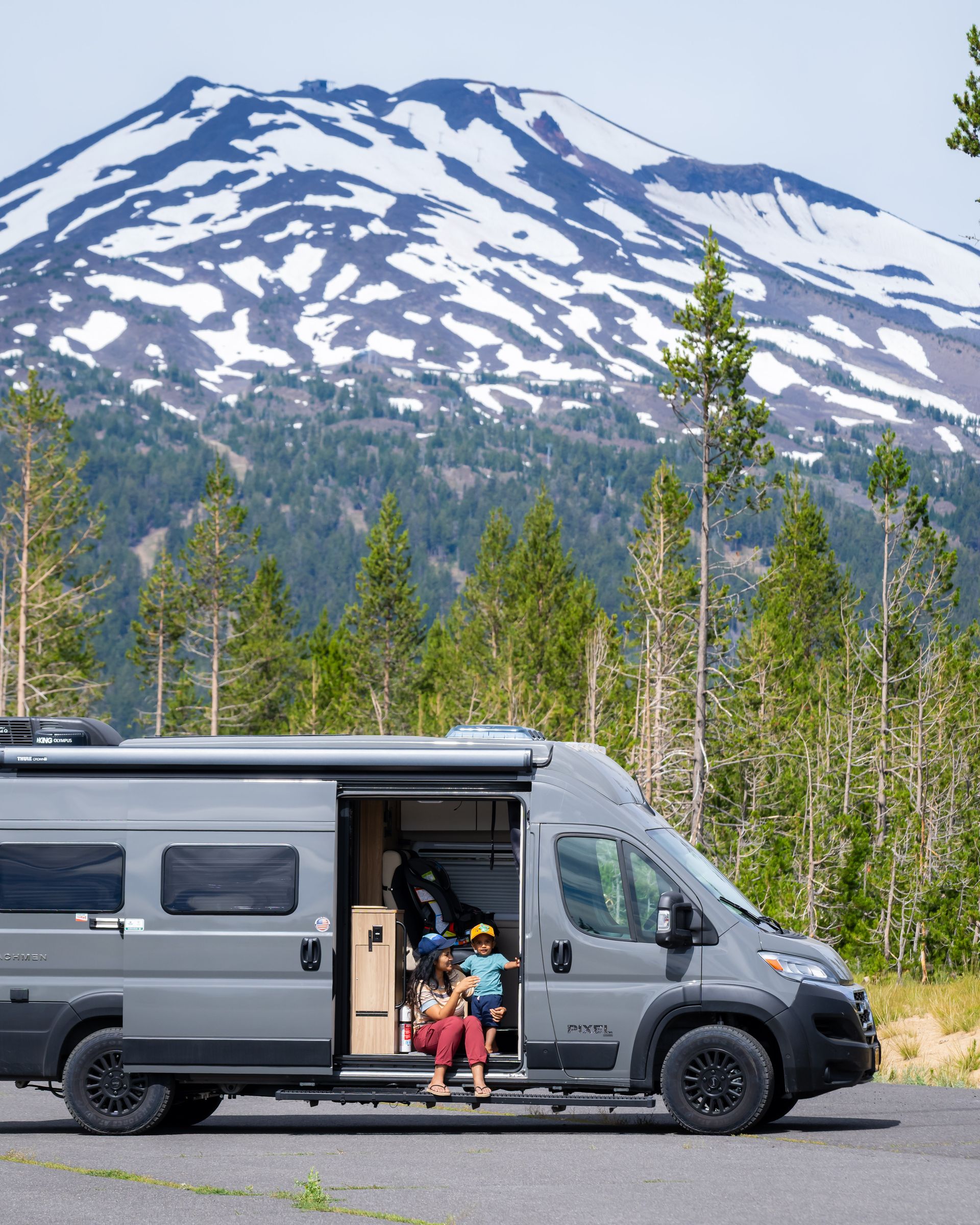 Camper van parked by snowy mountain, with people sitting in the open side door.