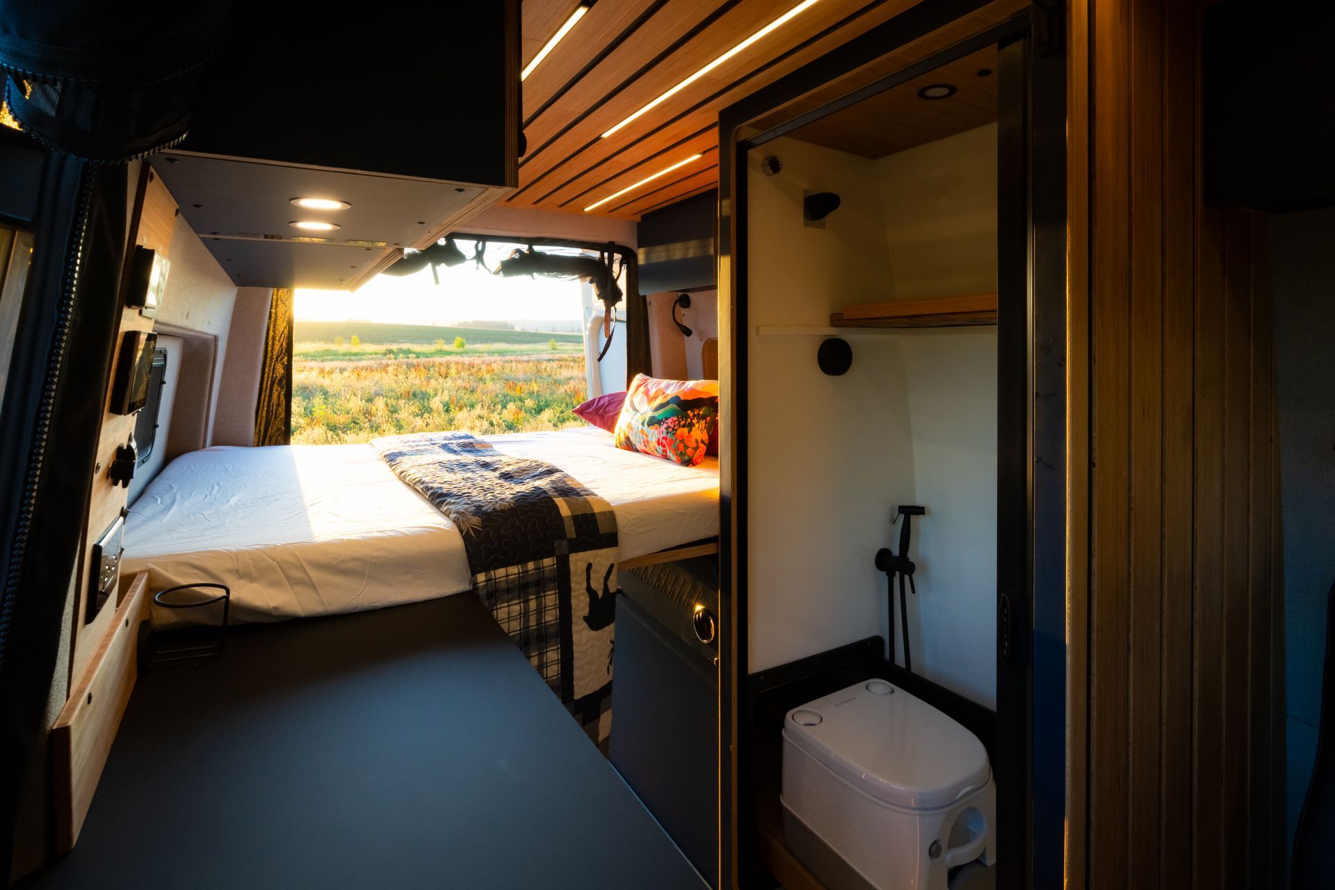 Interior of a camper van with a bed, kitchenette, and open doors overlooking a sunny landscape