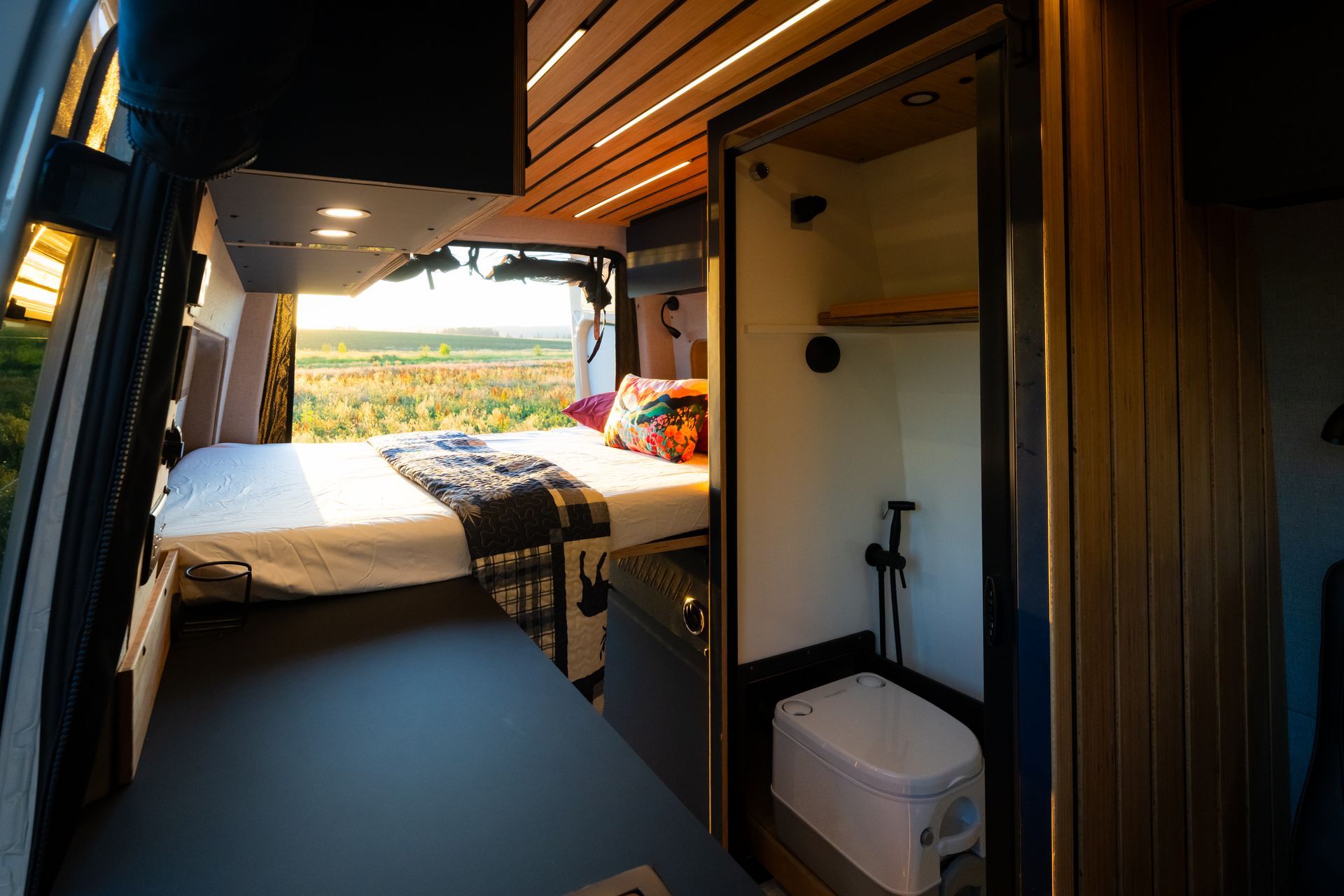 Compact camper van interior with bed, kitchenette, and open doors overlooking a scenic landscape.