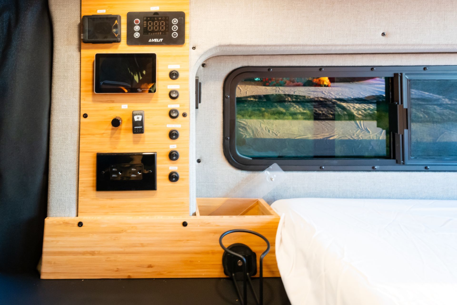 Wood-paneled van interior with bed, control panel, and window beside a small wooden desk.