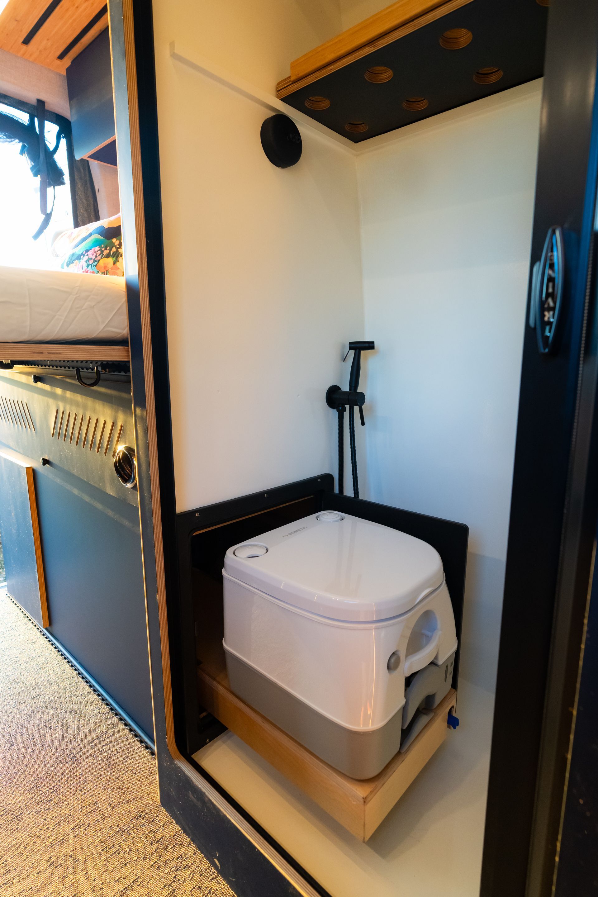 Compact white portable toilet in a narrow camper bathroom, with a handheld shower and dark wood trim.